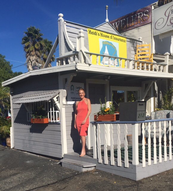 Heidi Bounds standing on the porch of Heidi's House of Insurance — a gray cottage office with white trim, flower boxes, and her name on the sign.